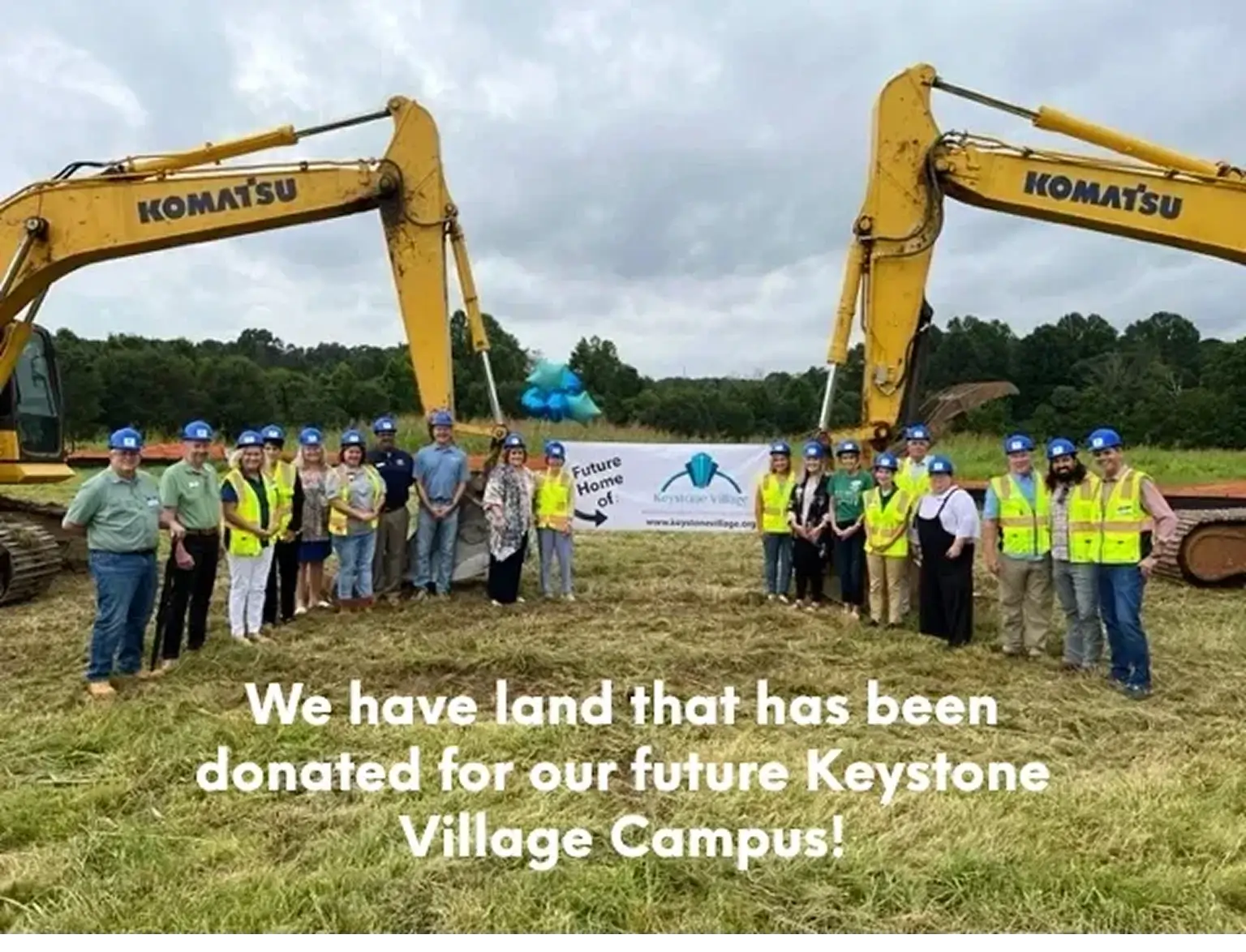 A large group of people wearing blue hard hats and neon safety vests stands in a grassy field for a groundbreaking ceremony. They are positioned between two large yellow Komatsu excavators and hold a white banner that reads "Future Home of: Keystone Village".